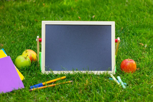 black blank chalkboard on the grass in the yard close-up, next to apples and school supplies