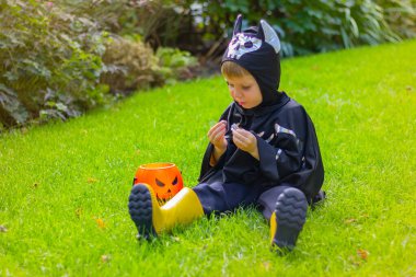 Yarasa iskeleti içinde cadılar bayramı kostümü giymiş küçük bir çocuk yeşil çimlerin üzerinde oturmuş çikolata tatlısı yiyor.
