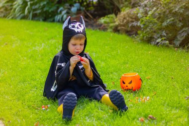 Siyah yarasa iskeleti içinde cadılar bayramı kostümü giymiş çocuk çimenlerde oturmuş çikolatalı şekerlemeler yiyor.
