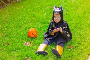 Yarasa iskeleti cadılar bayramı kostümü giymiş şirin çocuk ve sarı çizmeler çimenlerde oturmuş çikolata yiyor.