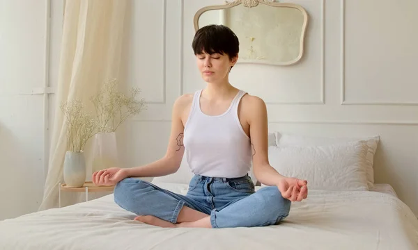 Genç bir kadın yatak odasındaki yatakta otururken meditasyon yapıyor. Sakin kadın derin nefes alıyor ve gözleri kapalı dinleniyor. Stres ve huzur yok..