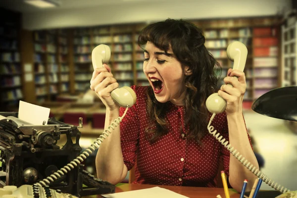 Mladá žena s červenými šaty zoufalá se dvěma telefony v obou rukou v úřadu, 1960 — Stock fotografie