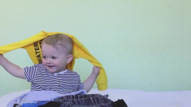 A cute baby is holding a huge stack of baby clothes and helping the mother with the housework. The boy is cleaning up the house. The child scatters things. Copy space- fun concept, organizing storage — Stok video