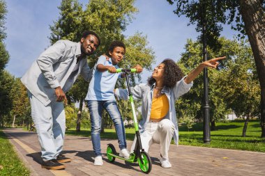 Güzel, mutlu, Afro-Amerikan aile parkta kaynaşıyor, siyahi aile dışarıda eğleniyor, aileler oğluna scooter sürmeyi öğretiyor.