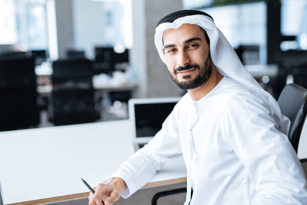 Handsome man with dish dasha working in his business office of Dubai. Portraits of a successful businessman in traditional emirates white dress. Concept about middle eastern cultures, lifestyle and professional occupations