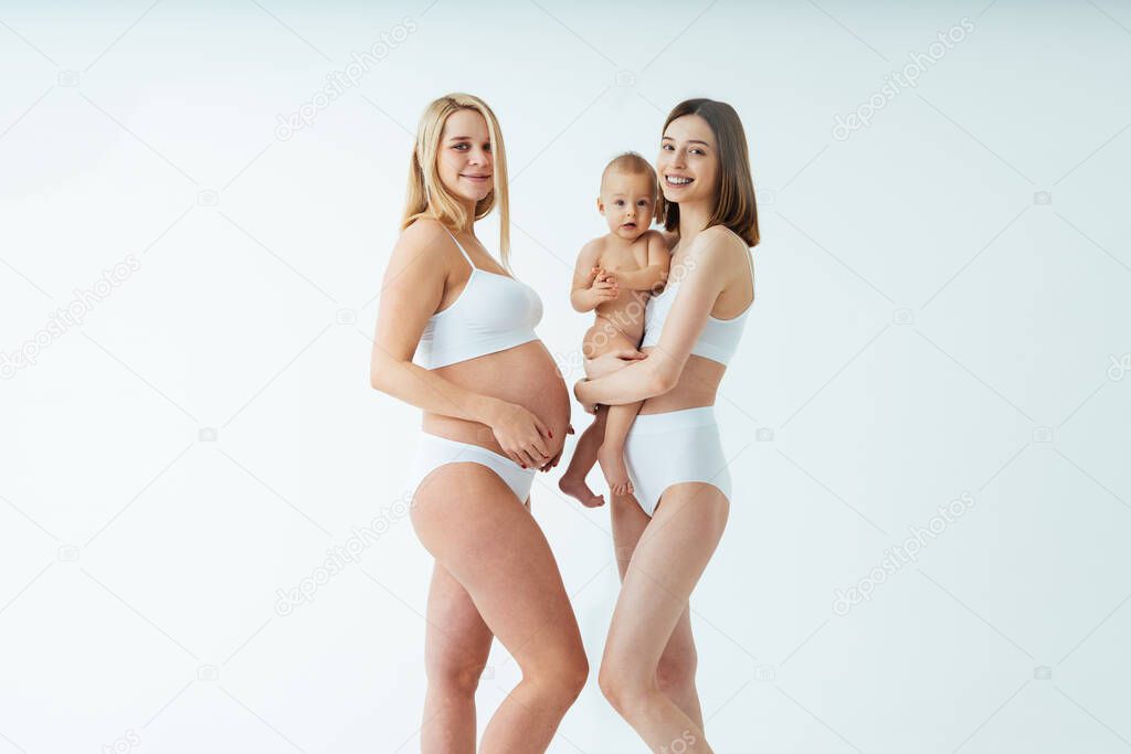 Happy pregnant woman with big belly and beautiful mother with baby on colored background -  Young women wearing underwear expecting a baby - Pregnancy, motherhood, people and expectation concept