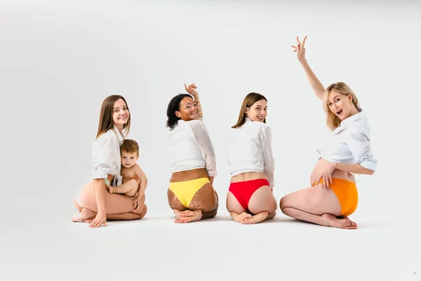 Happy pregnant women with big belly and beautiful mothers with babies in studio - Multiracial group of pregnant women and moms with childs wearing underwear - Pregnancy, motherhood, people, body positive and body acceptance concept