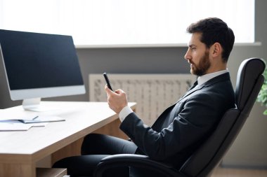 Ofisteki bilgisayar masasında oturan şık takım elbiseli bir iş adamı cep telefonunda uygulama kullanan yetişkin bir adam, iş ve teknoloji kavramları.