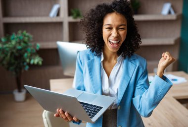 Zarif elbiseli güzel bir iş kadını bir iş stüdyosunda duruyor ve bilgisayar laptopunu online bir video konferansı için kullanıyor. Bir iş toplantısında Afrikalı Amerikalı kadın portresi, iş ve teknoloji konseptleri.