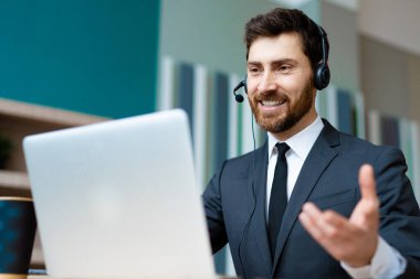 Zarif takım elbiseli bir işadamı ofiste bilgisayar masasında oturup bir video konferansına katılıyor - bir müşteri hizmetleri şirketi için internet ve dizüstü bilgisayarda çalışan yetişkin bir adam, iş, pazarlama, satış ekibi ve teknoloji konseptleri