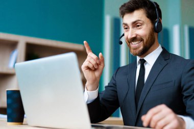 Zarif takım elbiseli bir işadamı ofiste bilgisayar masasında oturup bir video konferansına katılıyor - bir müşteri hizmetleri şirketi için internet ve dizüstü bilgisayarda çalışan yetişkin bir adam, iş, pazarlama, satış ekibi ve teknoloji konseptleri