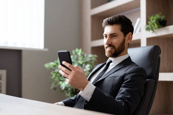 オフィスのコンピュータデスクに座ってエレガントなスーツを着たビジネスマン 携帯電話でアプリを使用して大人の男オンライン ビジネスと技術に関する概念 — ストック写真