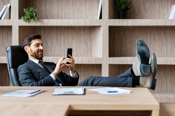 オフィスのコンピュータデスクに座ってエレガントなスーツを着たビジネスマン 携帯電話でアプリを使用して大人の男オンライン ビジネスと技術に関する概念 — ストック写真