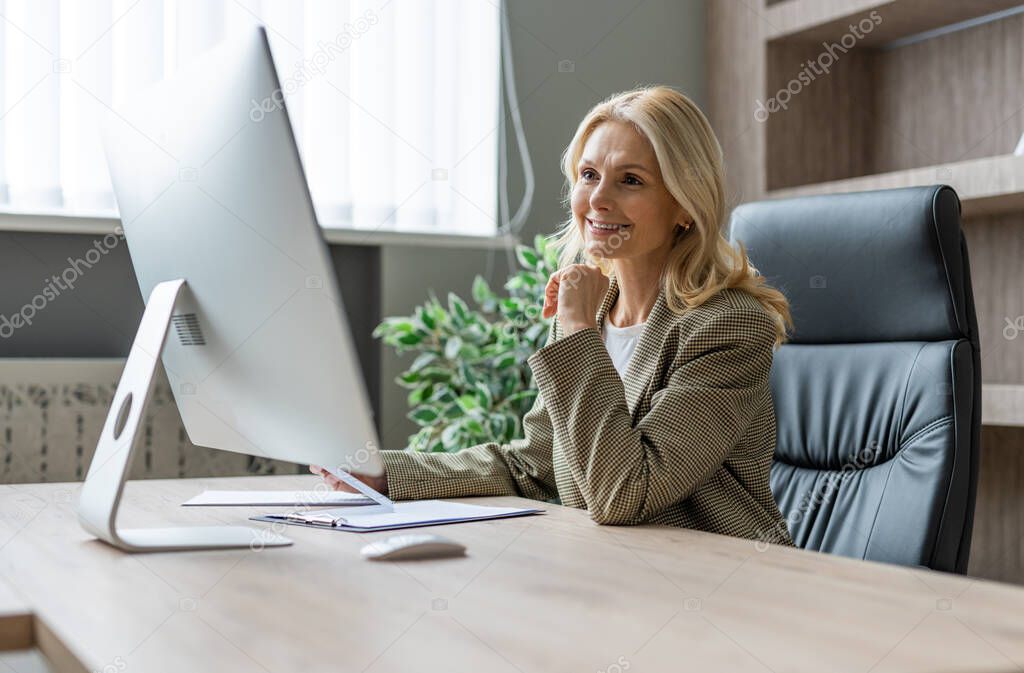 Beautiful mature adult businesswoman with elegant dress sitting at computer desk in the office - Senior woman working at pc online, concepts about business and technology