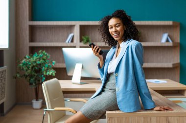 Zarif elbiseli güzel bir iş kadını bir iş stüdyosunda duruyor ve cep telefonu uygulaması kullanıyor. İş görüşmesinde Afro-Amerikan kadın portresi, iş ve teknoloji konseptleri.