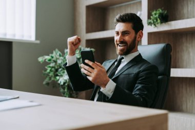 Ofisteki bilgisayar masasında oturan şık takım elbiseli bir iş adamı cep telefonunda uygulama kullanan yetişkin bir adam, iş ve teknoloji kavramları.