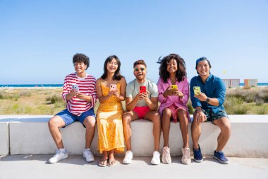 Multiethnic group of young happy friends bonding outside, having fun on summertime vacation - Multicultural cheerful people with summer clothes using mobile phones, concepts about youth, friendship and communication technology