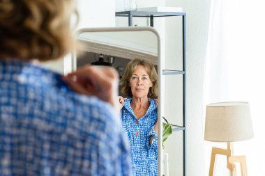 Beautiful mature senior woman at home, domestic life and leisure moments - 50-60 years old pretty female adult looking at herself in the mirror