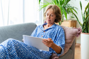Beautiful mature senior woman at home, domestic life and leisure moments - 50-60 years old pretty female adult relaxing on the sofa and using computer tablet