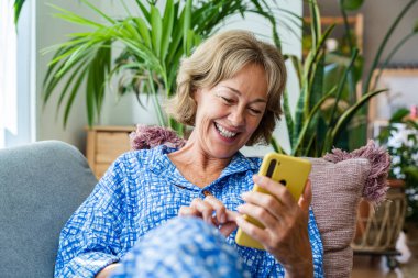 Beautiful mature senior woman at home, domestic life and leisure moments - 50-60 years old pretty female adult relaxing on the sofa and using smartphone