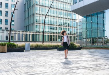 Beautiful hispanic businesswoman with elegant suit walking in the business centre- Adult female with business suit and holding mobile phone portrait outdoors