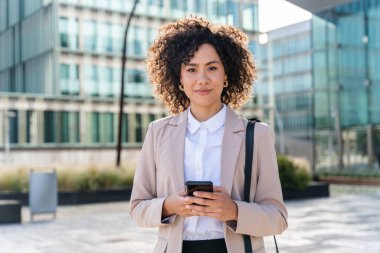 Beautiful hispanic businesswoman with elegant suit walking in the business centre- Adult female with business suit and holding mobile phone portrait outdoors