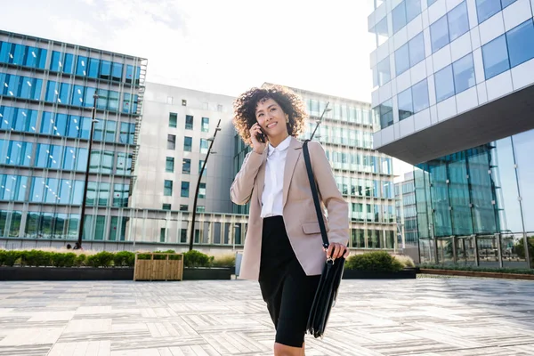 Beautiful hispanic businesswoman with elegant suit walking in the business centre- Adult female with business suit and holding mobile phone portrait outdoors