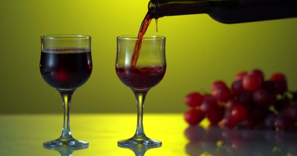 El vino tinto forma una ola hermosa. Vino que vierte en copa de vino en el fondo dramático. Primer plano de tiro. Una foto publicitaria. Uva fresca en la mesa reflectante. Fondo de color amarillo. Dos vasos. Holoday. — Vídeos de Stock