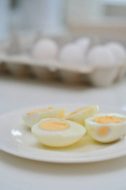 Mutfak masasında haşlanmış yumurta, sağlıklı kahvaltı ve protein zengini diyet. Tabakta yumurta, yanında bıçak, havadar ve beyaz arka plan, yeşil bitkiler ve masadan yansıyan ışık..