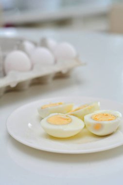 Mutfak masasında haşlanmış yumurta, sağlıklı kahvaltı ve protein zengini diyet. Tabakta yumurta, yanında bıçak, havadar ve beyaz arka plan, yeşil bitkiler ve masadan yansıyan ışık..