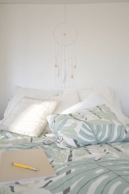Duvarda asılı bir yatak odasında düş kapanı ya da makrome. Bohem, uyumlu bir hayat ve iyi ruhlarla tatlı rüyalar. Boho şıklığı, etnik tılsım ve sembol, konforlu beyaz iç tasarım. Yatağın üzerinde bir defter ve kalem var.