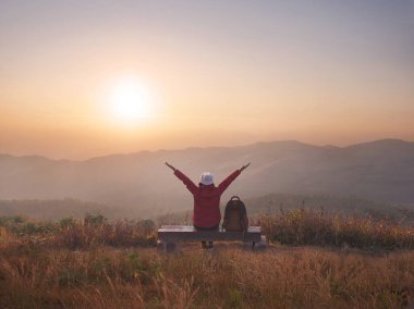 Arkaya bakan mutlu kadın sırt çantalı kişi tahta bankta oturur ve sabahın ilk ışıklarında dağın tepesinde kollarını kaldırır. Özgürlük yaşam tarzı insan kavramı.