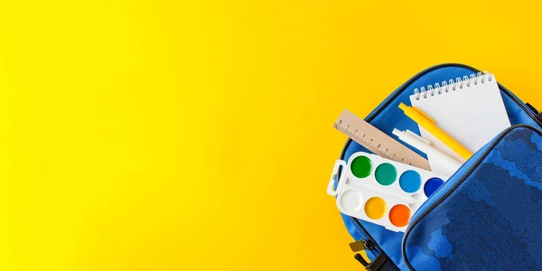 Conjunto de útiles escolares que sobresalen de la mochila azul en el banner de fondo amarillo — Foto de Stock