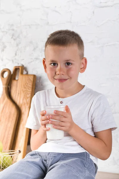 Um menino loiro bonito segurando um copo com leite e sorrindo. Alimentos saudáveis. — Fotografia de Stock