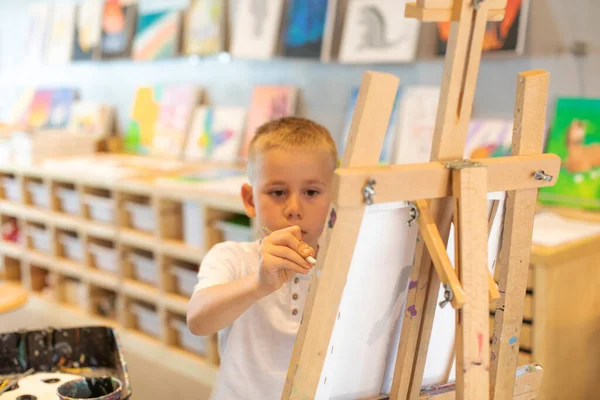 Un ragazzo biondo disegna su un cavalletto in una lezione di disegno. Il concetto di creatività dei bambini. — Foto Stock