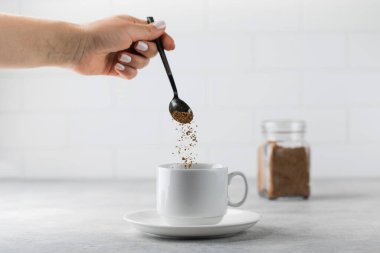A woman adds instant coffee to a white mug on grey stone table clipart