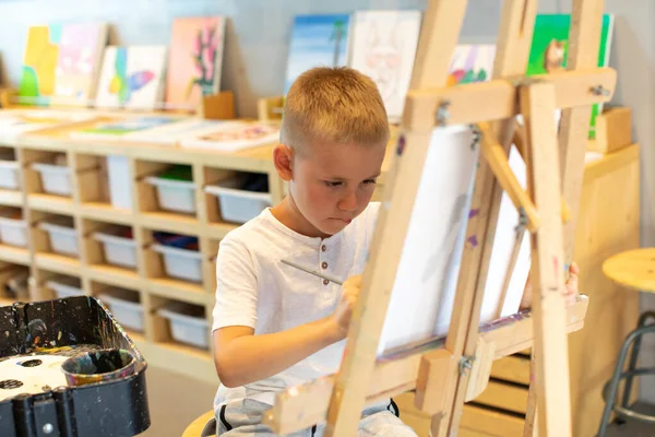 Un chico rubio dibuja en un caballete en una lección de dibujo. El concepto de creatividad infantil. —  Fotos de Stock