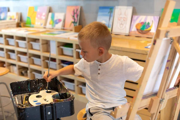 Un chico rubio mezcla pinturas mientras está sentado frente a un caballete en una lección de dibujo. El concepto de creatividad infantil. —  Fotos de Stock