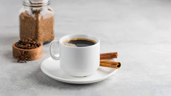 Café noir dans une tasse blanche avec des bâtons de cannelle sur une soucoupe sur une table grise. Espace de copie. — Photo