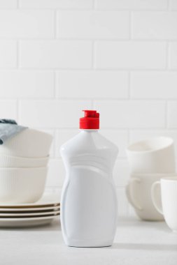 Beyaz fayanslı arka planı ve temiz bulaşıkları olan sıvı bulaşık deterjanıyla beyaz plastik bir şişe..
