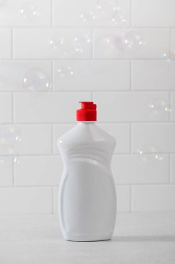 Beyaz plastik bir şişe, sıvı bulaşık deterjanı ve baloncuklu beyaz fayanslı bir arka plan. 