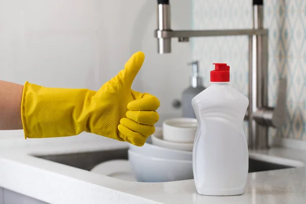 Biała butelka z żelem do zmywania naczyń i dłonią w żółtej gumowej rękawiczce macha kciukiem na zlewie z naczyniami. — Zdjęcie stockowe