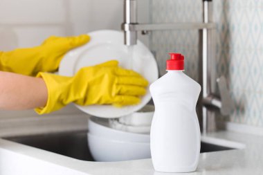 Bir ev hanımının arka planında bulaşık jeli olan beyaz bir şişe sarı lastik eldivenlerle lavaboda bulaşıkları yıkıyor..