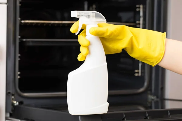 El limpiador sostiene una maqueta de una botella de detergente para horno delante del horno en un guante amarillo para el hogar. — Foto de Stock