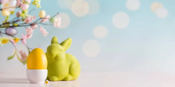 Easter banner with a yellow egg and a green rabbit on a blue background with a bokeh and a branch of flowers. Copy space — Stock Photo, Image