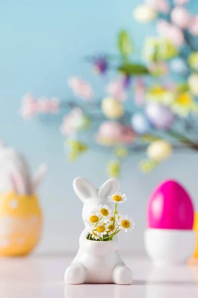 Tarjeta de Pascua. Un conejito blanco con un ramo de margaritas sobre un fondo azul y huevos de colores. — Foto de Stock