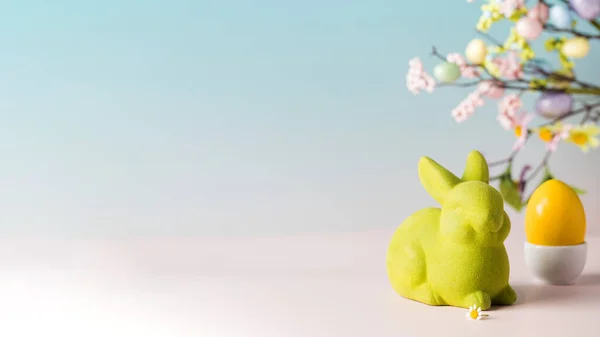 Easter banner with a yellow egg and a green rabbit on a blue background with a branch of flowers. Copy space — Stock Photo, Image