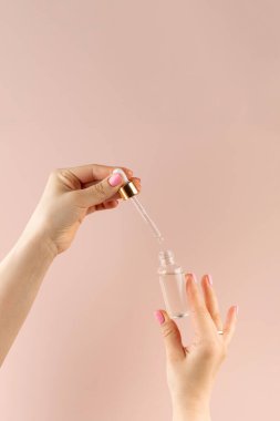 Kadın elleri, açık pembe arka planda özen ürünü olan bir damlanın içinde pipetle yağ damlatıyor. Bakım kozmetikleri.