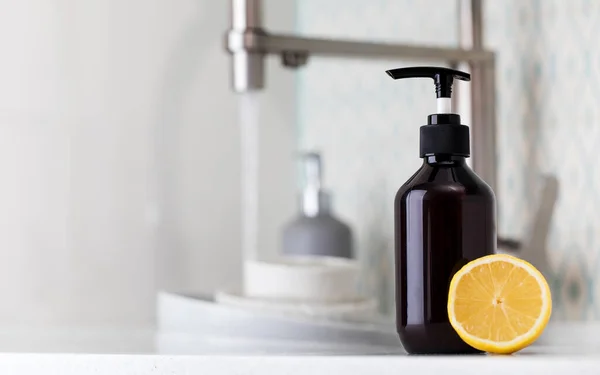 Botol dengan ramah lingkungan deterjen cuci piring dengan lemon di latar belakang wastafel dengan piring kotor. — Stok Foto