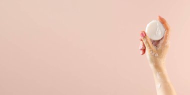 Bir kadının eli açık pembe arka planda yuvarlak bir sabunu tutar. Pankart